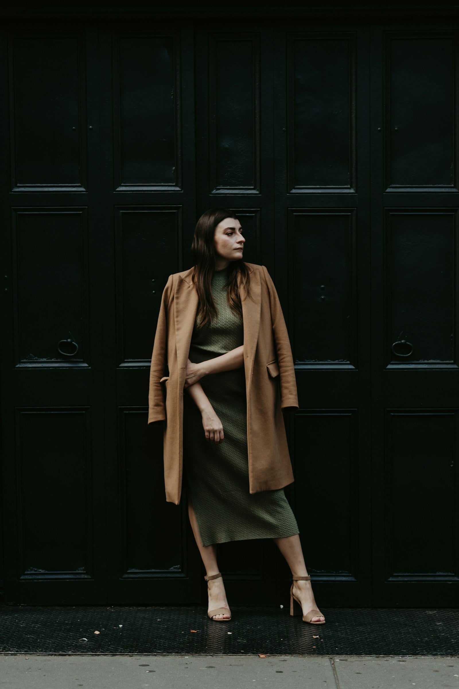 Woman in an olive midi dress and camel coat against a dark doorway