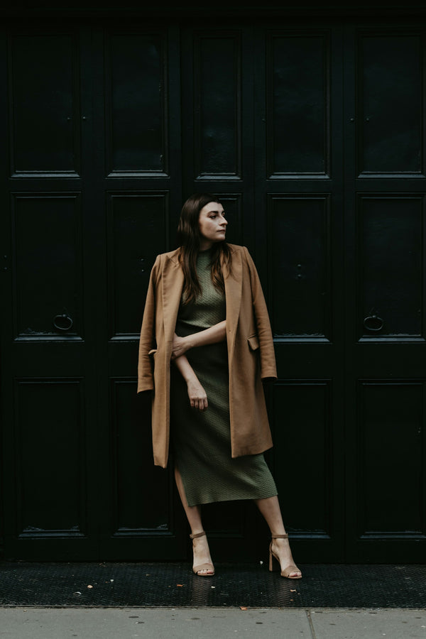 Woman in an olive midi dress and camel coat against a dark doorway